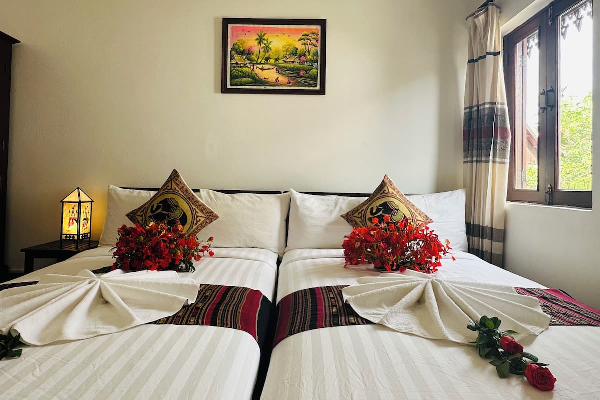 Habitación de hotel con una cama que tiene dos almohadas con diseños de elefantes, un cuadro de un paisaje en la pared y una ventana con cortinas.