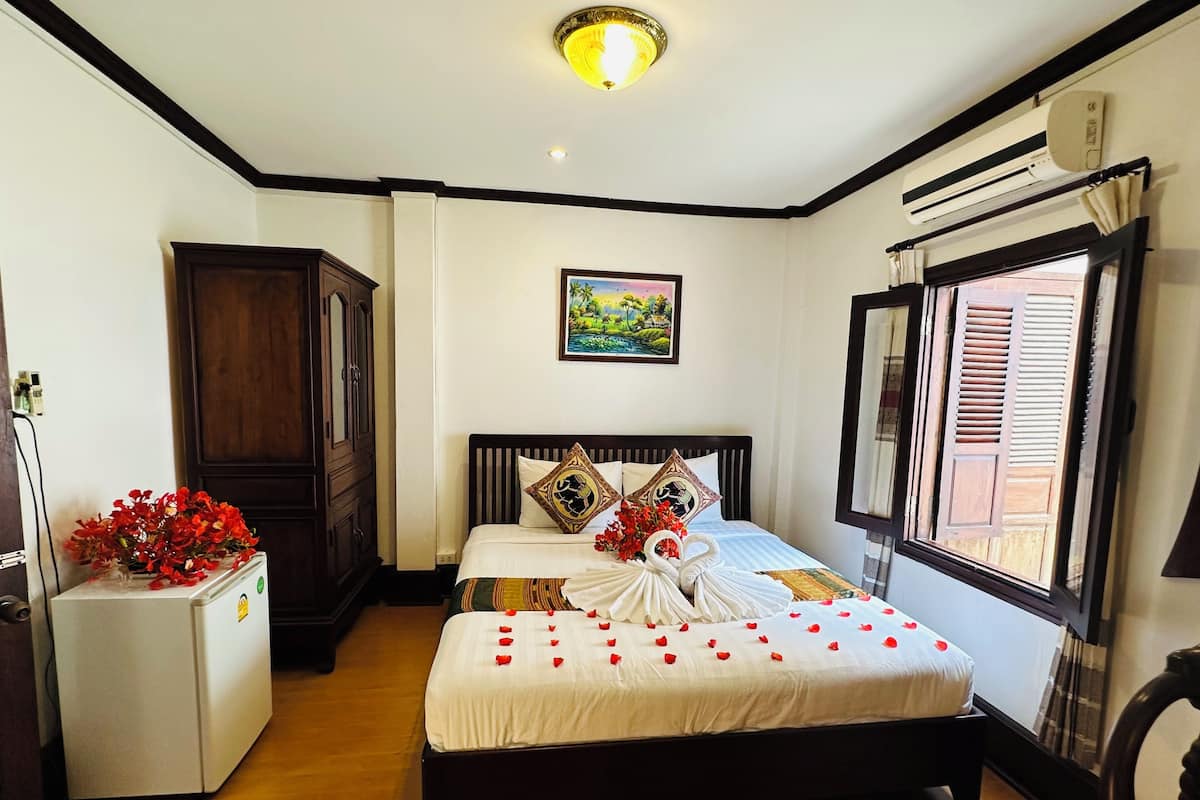 Habitación de hotel con piso de madera, cama con ropa blanca y almohadas decorativas, refrigerador blanco pequeño, aire acondicionado montado en la pared y ventana con persianas de madera.