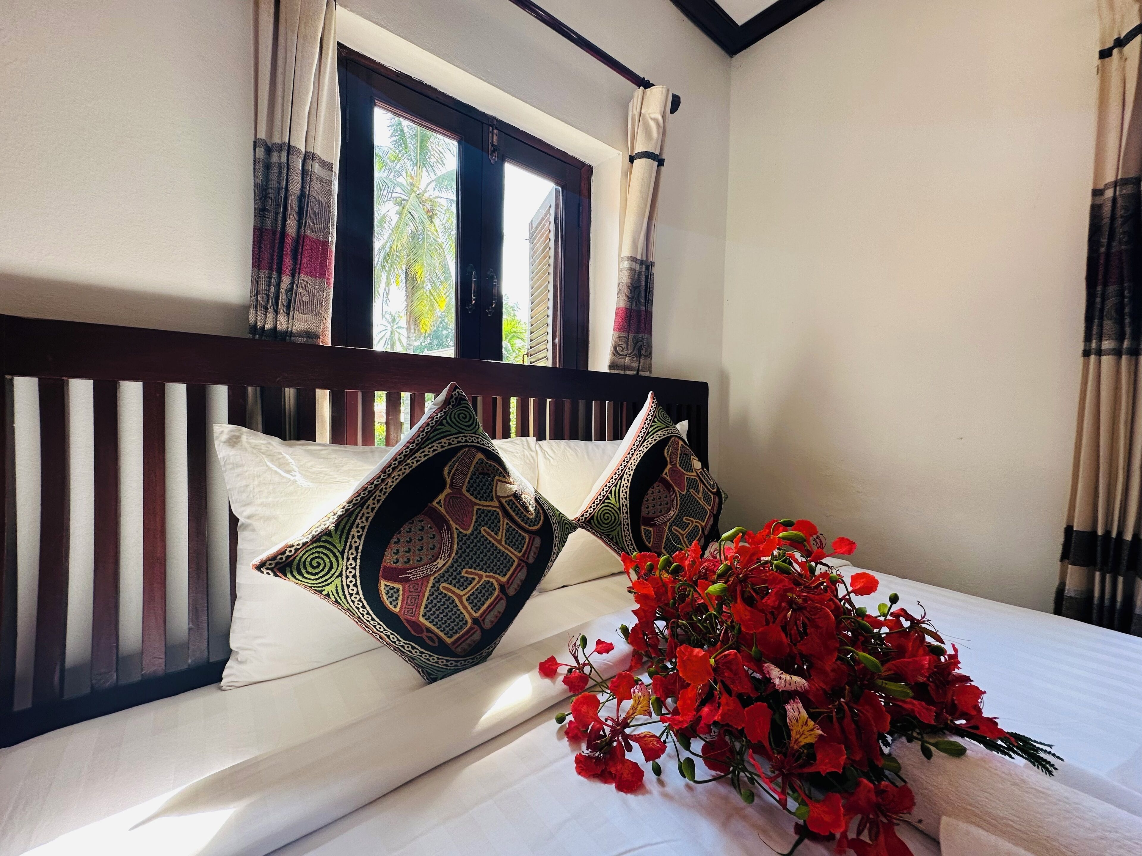 A bed with two decorative pillows, a window with curtains, and a bouquet of red flowers on the bed.