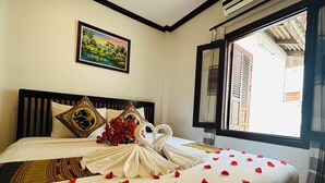 A neatly made bed with a decorative swan and rose petals, a framed landscape painting, and a window with shutters. - Thatsaphone Hotel (Luang Prabang)
