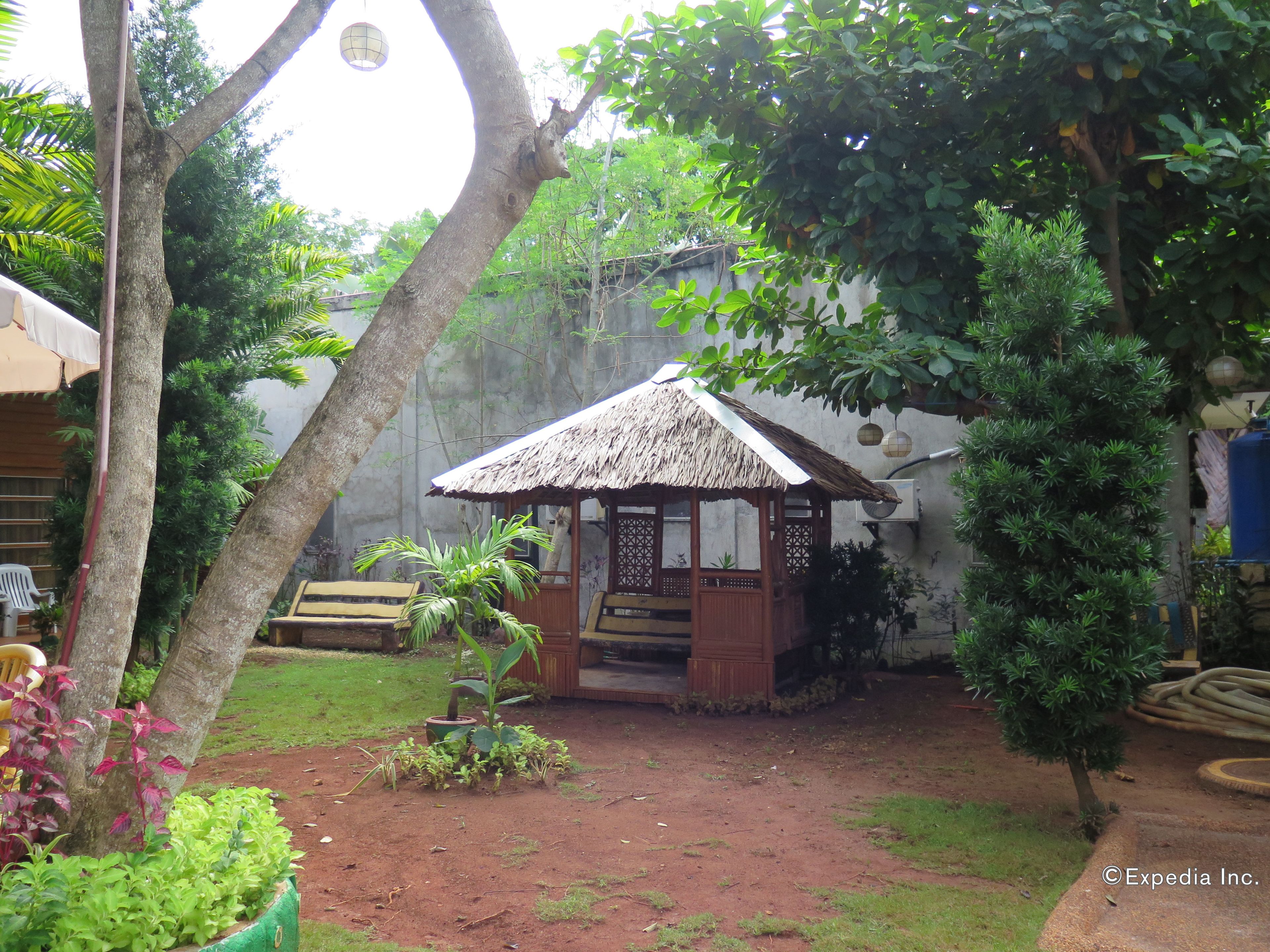 A thatched-roof gazebo in a garden with a bench, surrounded by various plants and trees.