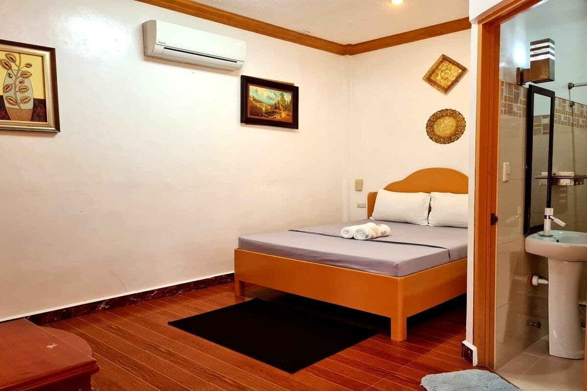 A bedroom with a bed, a bedside table, an air conditioner, a framed picture, and a bathroom with a sink and shower.