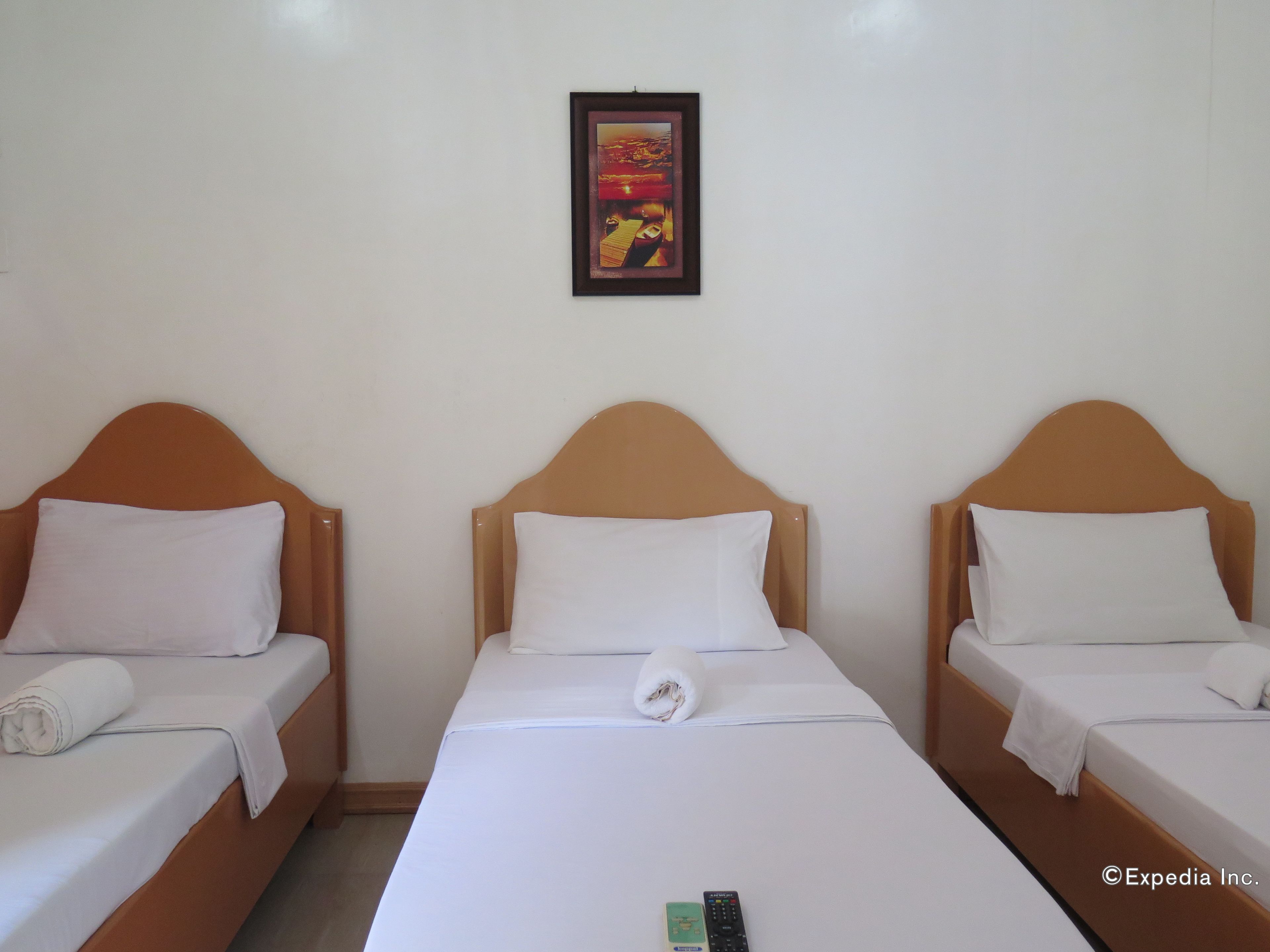 Une chambre d’hôtel avec trois lits simples, chacun doté d’une tête de lit en bois et de draps blancs. Un tableau encadré est accroché au mur, au-dessus des lits.