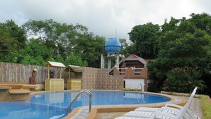 Una piscina con fuente de agua azul, rodeada por una valla de madera y un muro de ladrillo. Hay sillas de descanso blancas y una pequeña estructura de madera con techo de paja.