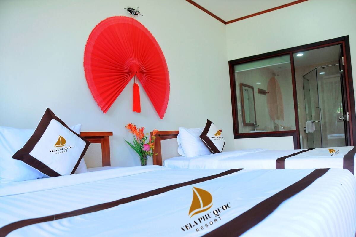 A hotel room with two beds, a red fan, a vase with flowers, and a glass shower door.