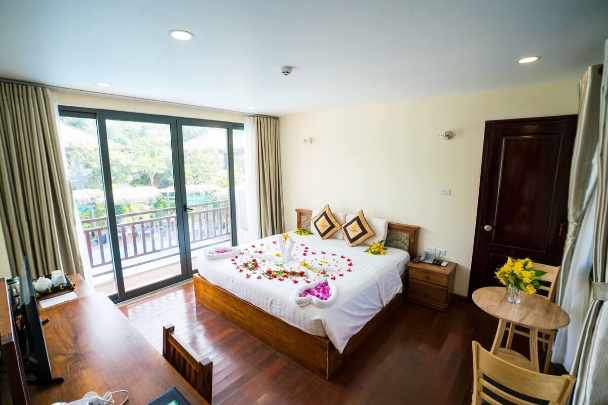 Quarto de hotel com uma cama grande, uma escrivaninha de madeira, uma cadeira, uma mesinha com flores e uma varanda com vista.