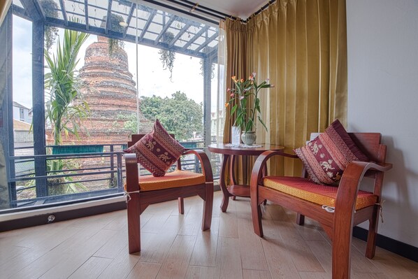 A room with two wooden chairs and a small table, a large window with a view of a temple and greenery, and a curtain on the window. - Chedi Home Chiang Mai (Chiang Mai)
