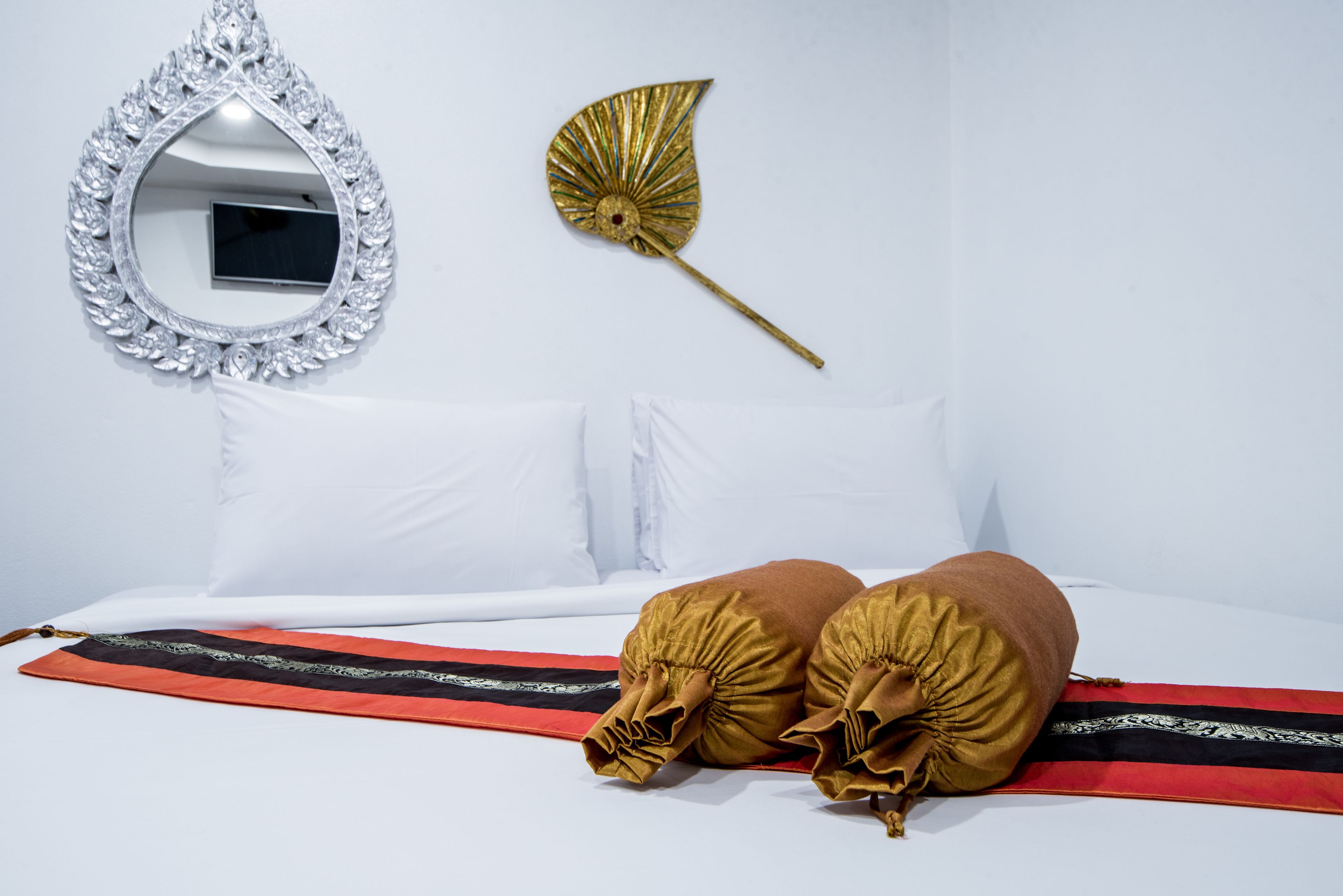 Una cama con almohadas blancas y una colcha a rayas rojas y negras, un abanico decorativo dorado y un espejo con marco plateado en la pared.