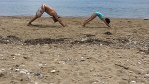 Duas pessoas praticando uma postura de yoga em uma praia arenosa, com um mar calmo e montanhas ao fundo.