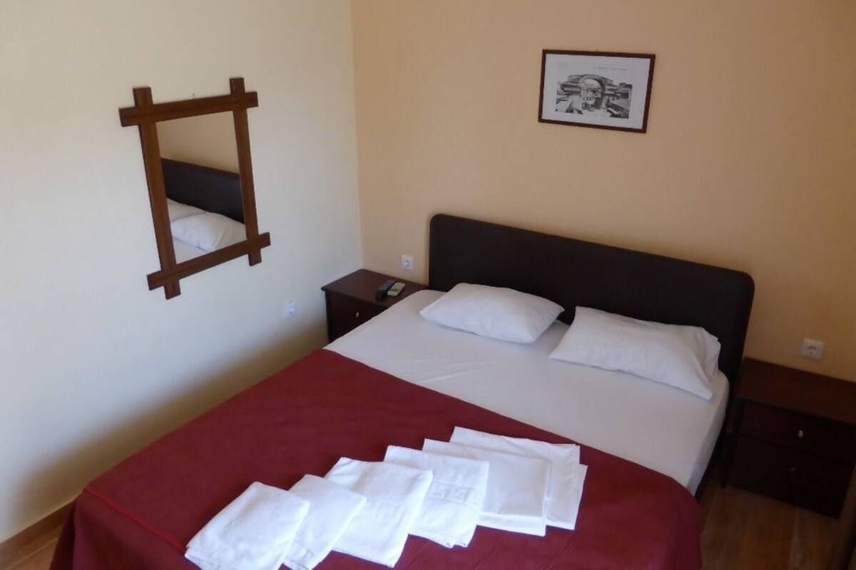 Una cama bien hecha con sábanas blancas y una manta roja, una mesita de noche de madera con un teléfono, un cuadro en la pared y un espejo a la izquierda.