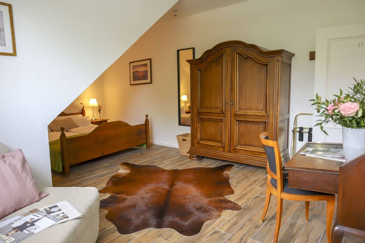A bedroom with a wooden bed, a large wardrobe, a desk with a chair, a leather rug, and a small potted plant.