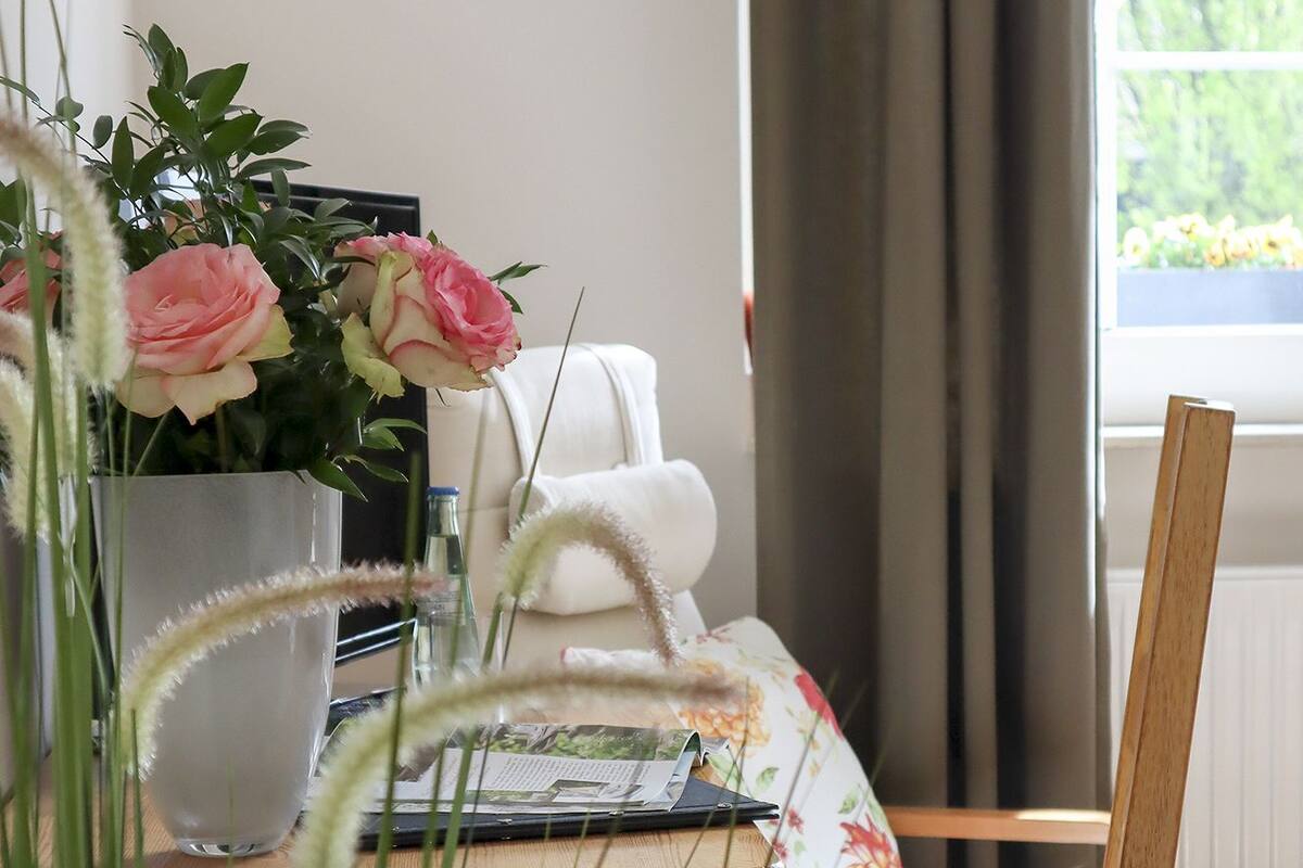 A wooden chair with a woven seat, a white cushion, and a floral-patterned pillow. In the background, there is a white chair, a window with a view of greenery, and a vase with pink flowers.