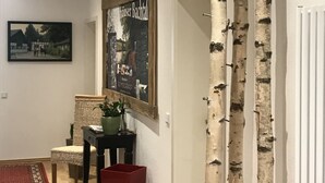 A hallway with a wooden floor, a red rug, a framed poster, a chair, a small table with a plant, and birch tree trunks against the wall. - Hotel Gut Moschenhof (Düsseldorf)