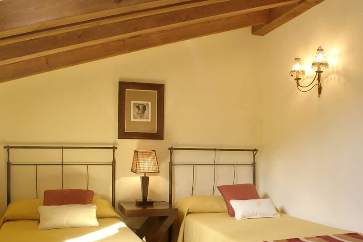 A room with two beds, wooden beams on the ceiling, a framed picture on the wall, and a wall-mounted lamp.
