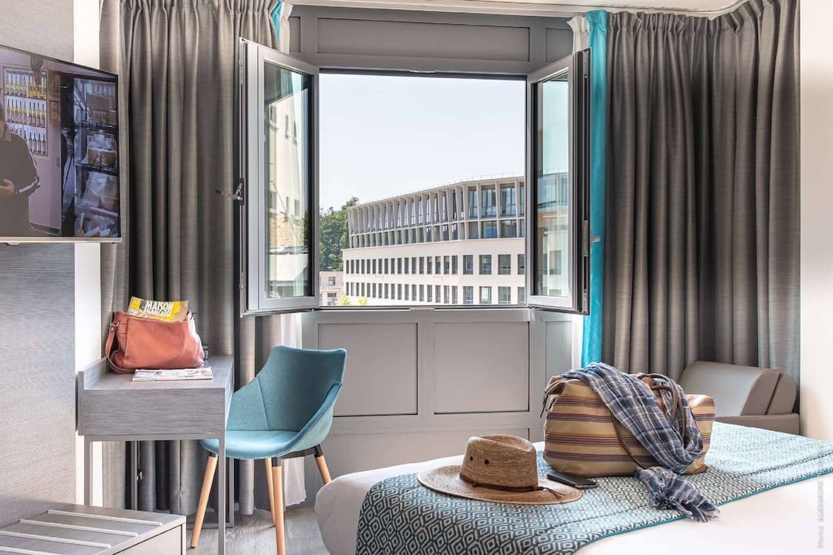 Habitación de hotel con un ventanal grande, un escritorio con una computadora portátil, una silla, una cama con colcha estampada y una maleta.