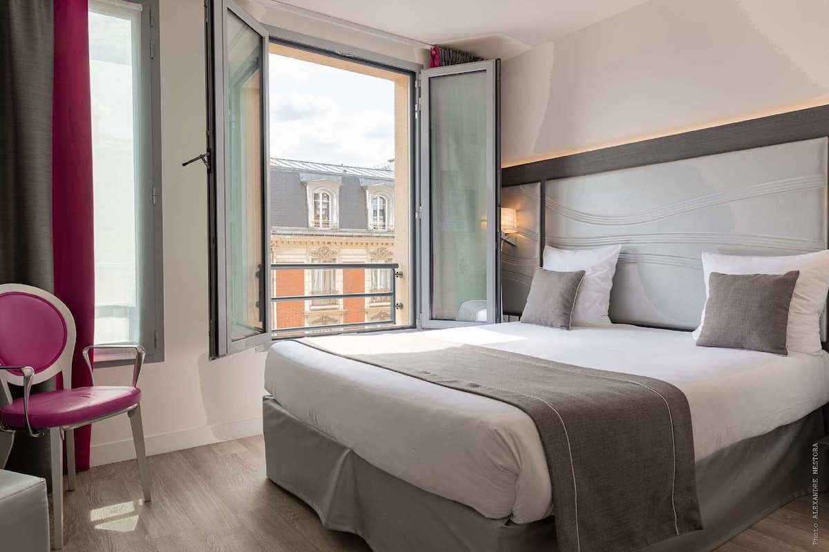 Habitación de hotel con cama, una silla, ventana con cortinas y vistas a edificios.