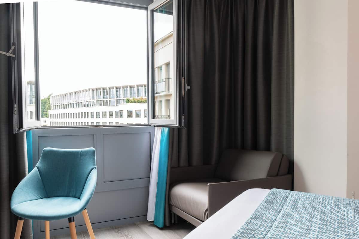 Una habitación de hotel moderna con una cama, una silla azul, una ventana con cortinas y vistas a un edificio.