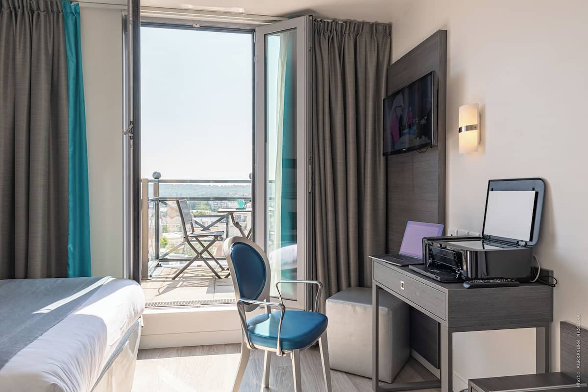 Habitación de hotel con una cama, un escritorio con una computadora portátil, una silla y vistas de la ciudad a través de un ventanal grande.