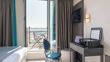 Habitación de hotel con una cama, un escritorio con una computadora portátil, una silla y vistas de la ciudad a través de un ventanal grande.