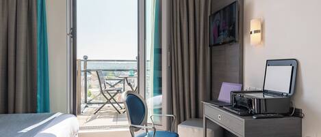 A hotel room with a bed, a desk with a laptop, a chair, and a view of the city through a large window.