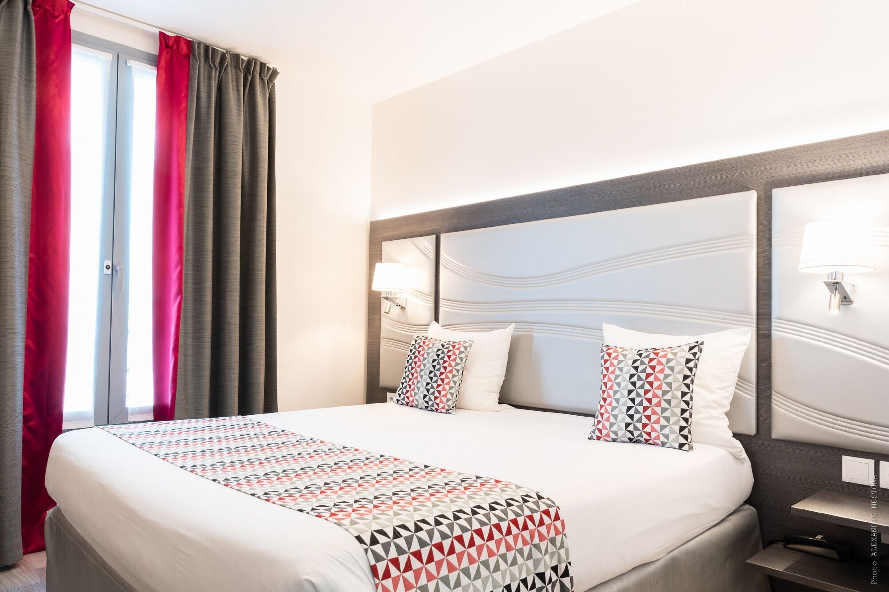 Una habitación de hotel con una cama grande, dos lámparas de noche y un ventanal con cortinas.