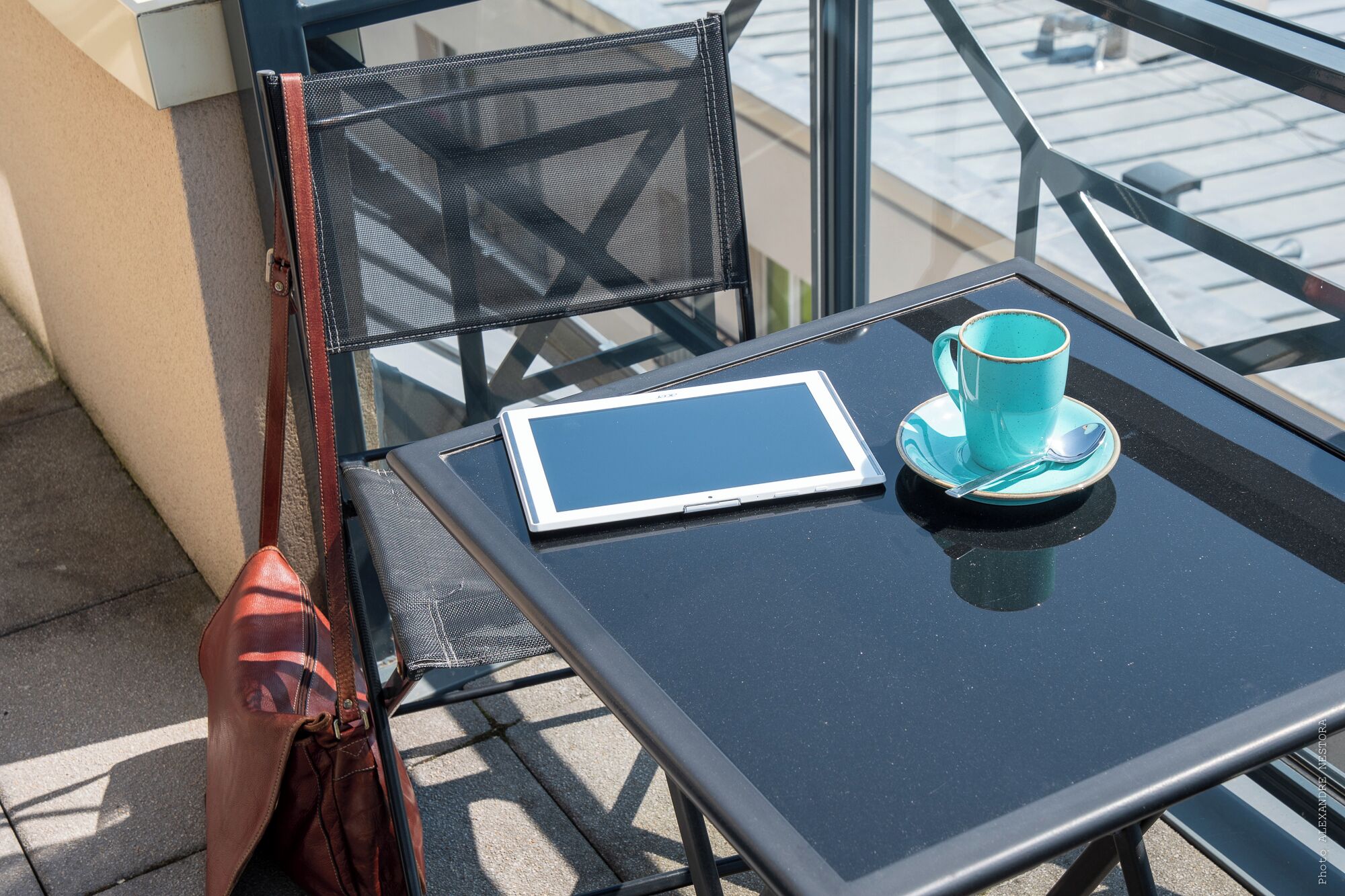 A tablet, a blue cup, and a brown bag on a balcony table.