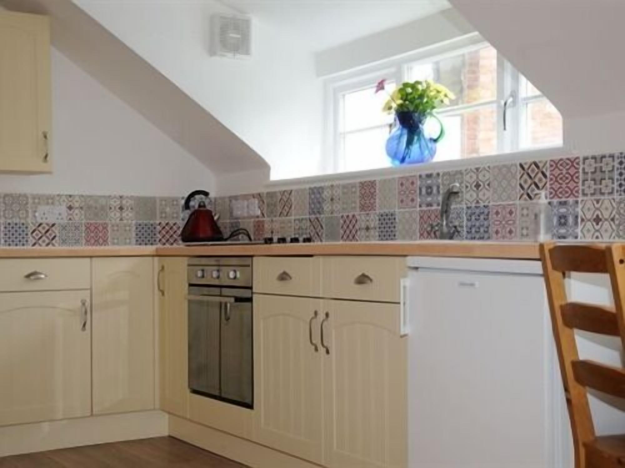 A kitchen with a tiled backsplash, a kettle, a refrigerator, and wooden cabinets.