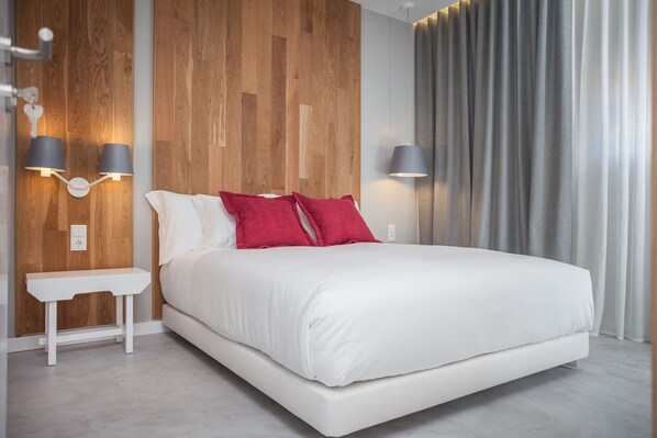 A modern bedroom with a wooden accent wall, a white bed, red pillows, and two wall-mounted lamps. - Vale d'Azenha Hotel and Residences (Alcobaça)