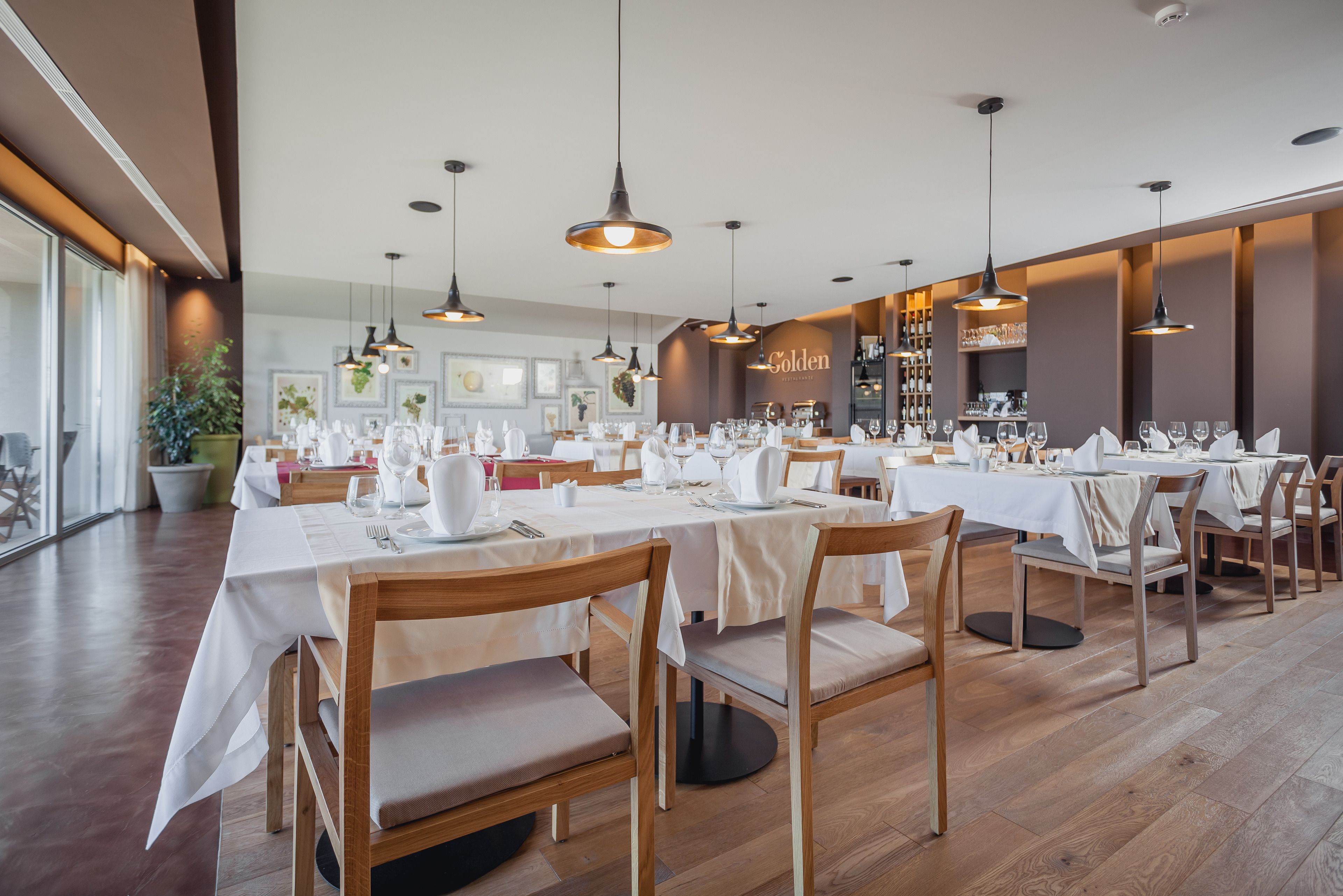 A restaurant with multiple tables set for dining, wooden chairs, pendant lighting, and a visible 'Golden' sign on the wall.