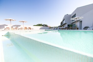 A swimming pool with lounge chairs and umbrellas, a modern building with large windows, and a clear sky. - Vale d'Azenha Hotel and Residences (Alcobaça)