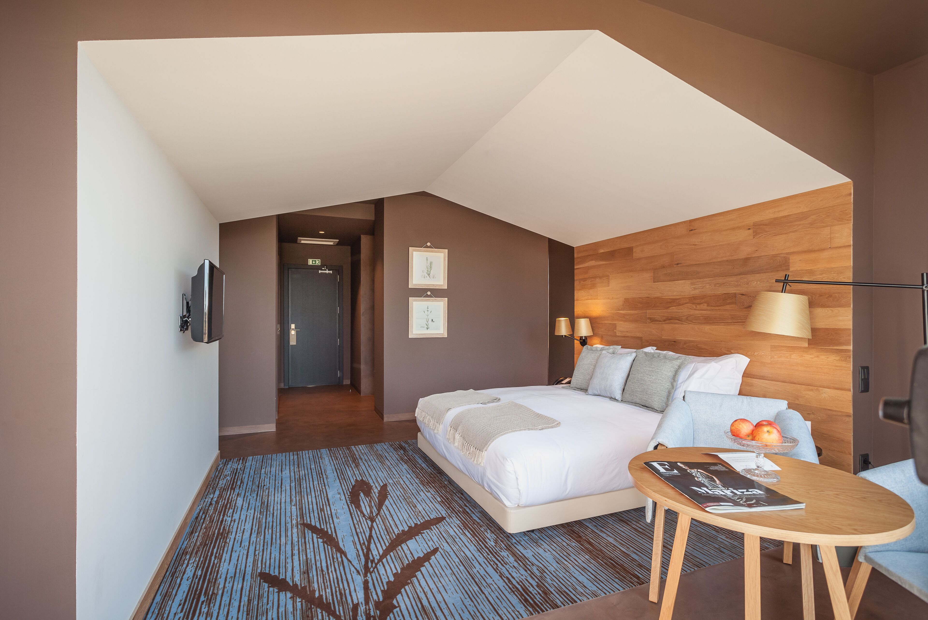 A modern hotel room with a bed, a small table, a TV, and a wooden accent wall.