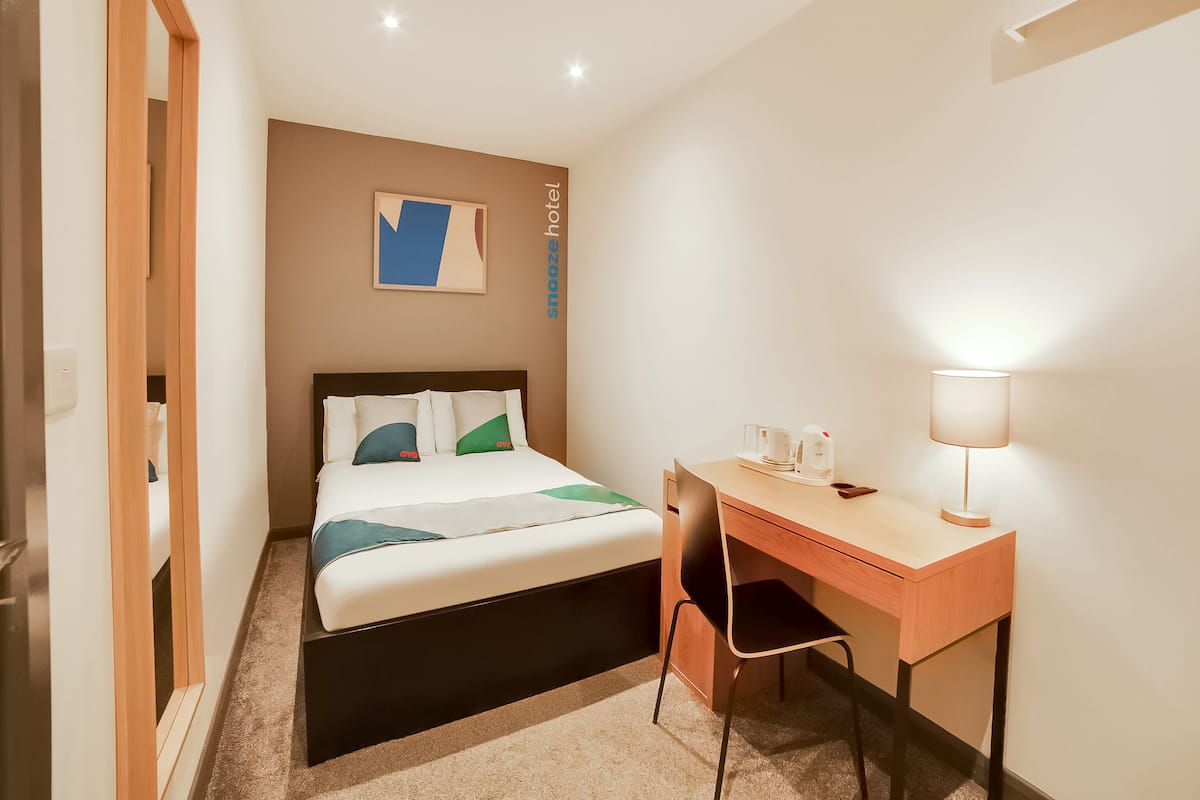 A hotel room with a bed, a desk, a chair, a lamp, and a painting on the wall.