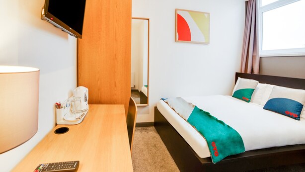 Habitación de hotel con cama, escritorio, televisor, cafetera y un cuadro en la pared.