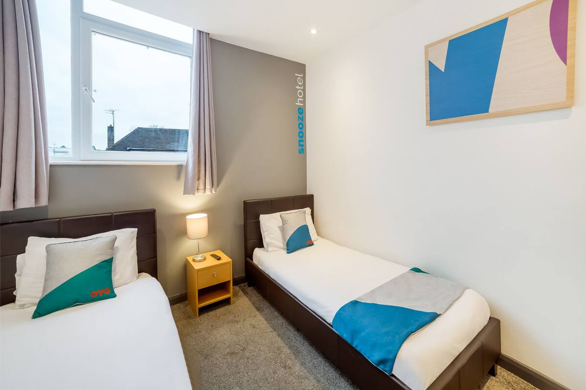 A hotel room with two single beds, a bedside table with a lamp, a window with curtains, and a wall-mounted artwork.