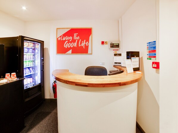 Un área de recepción con mostrador de madera, una máquina expendedora y un cartel en la pared que dice "Living The Good Life".