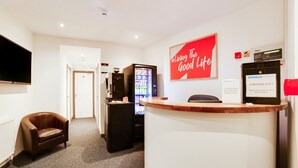 Zona de recepción de un hotel con un sillón de cuero marrón, un refrigerador, una máquina expendedora y un televisor en la pared.
