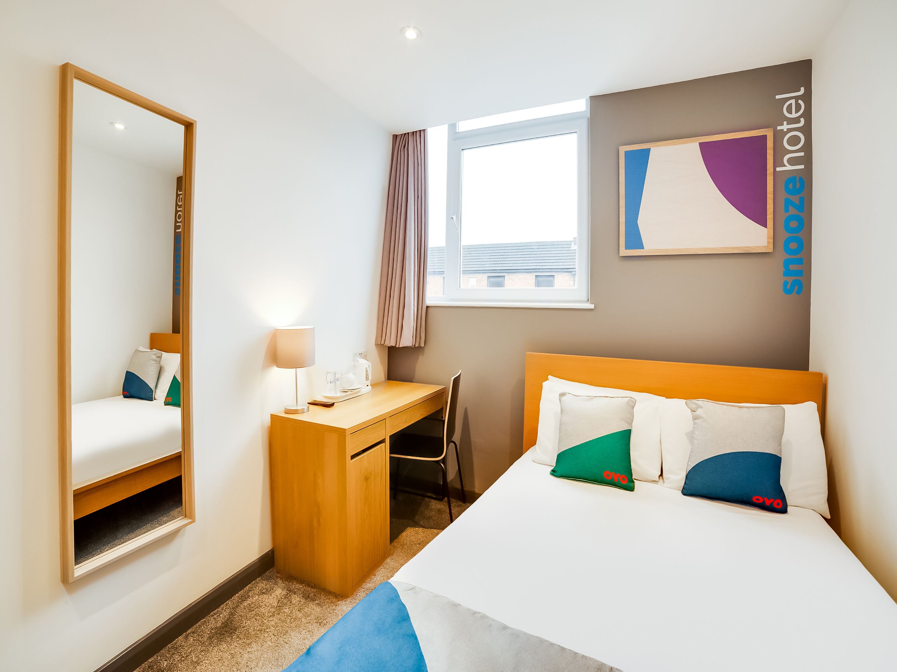 Una habitación de hotel con cama, escritorio, silla, espejo, ventana y una obra de arte colgada en la pared.