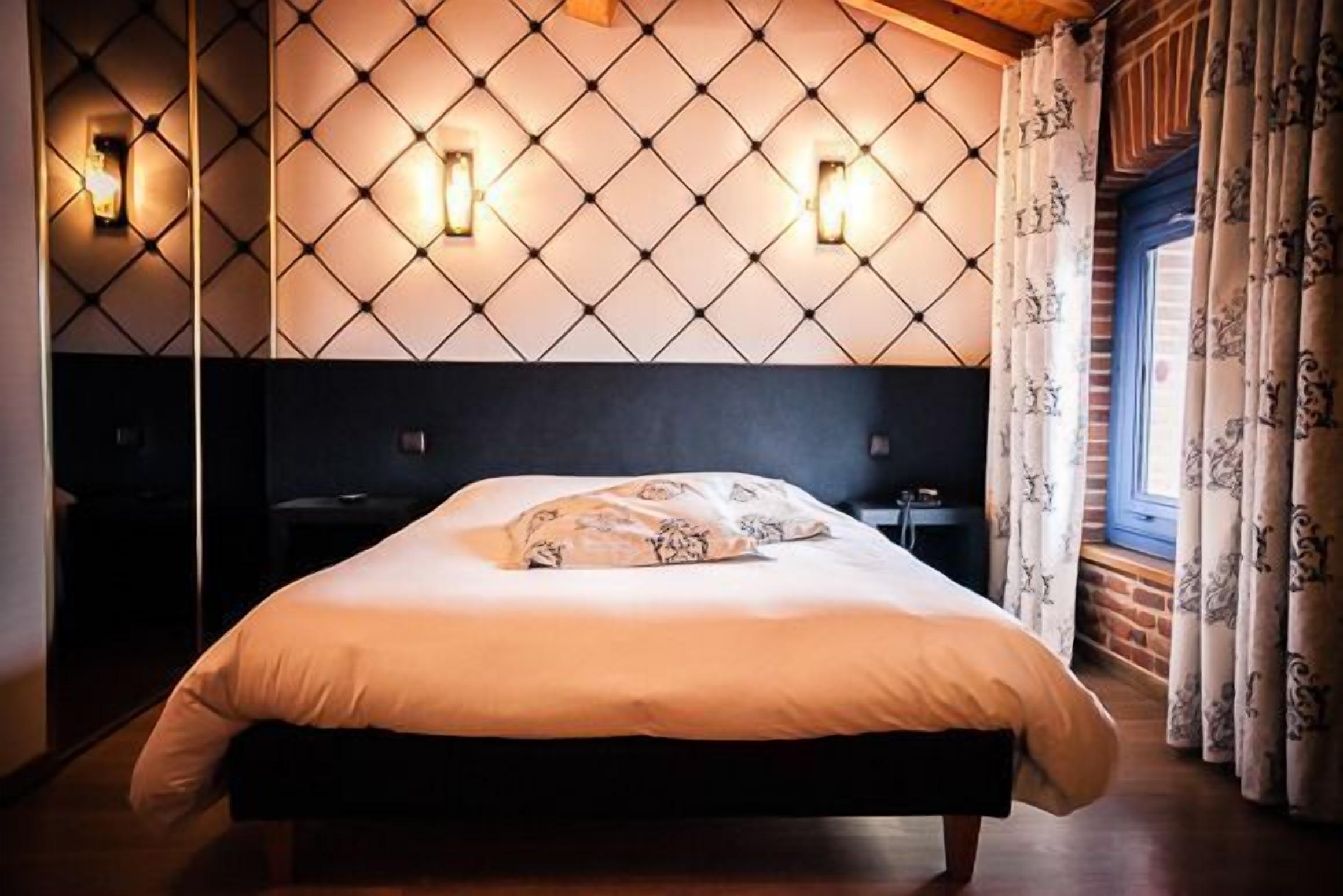 A neatly made bed with a patterned pillow in a room with a diamond-patterned headboard, two wall-mounted lights, and a window with curtains.