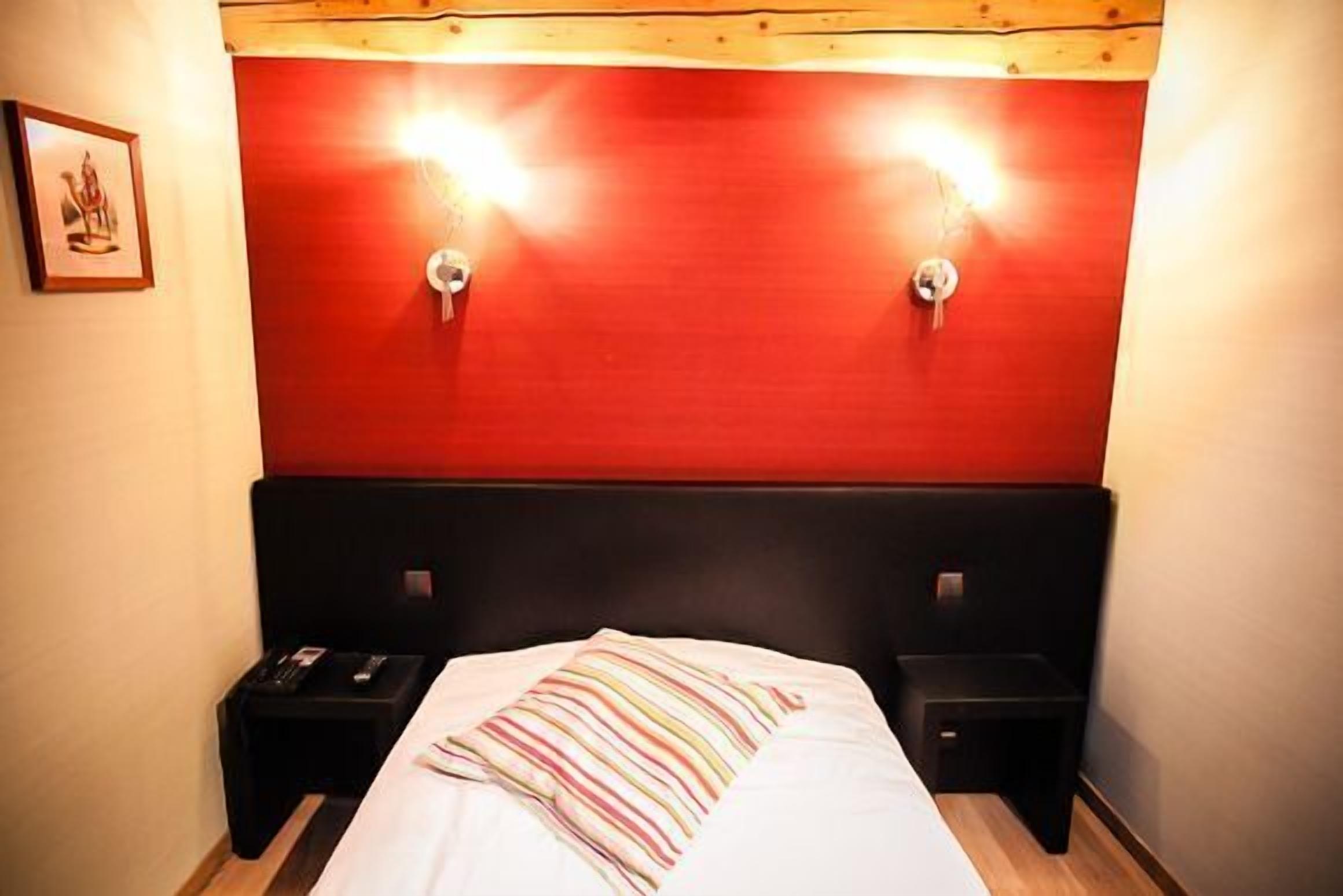 Quarto de hotel com cabeceira vermelha, duas mesinhas de cabeceira e uma cama com travesseiro listrado.