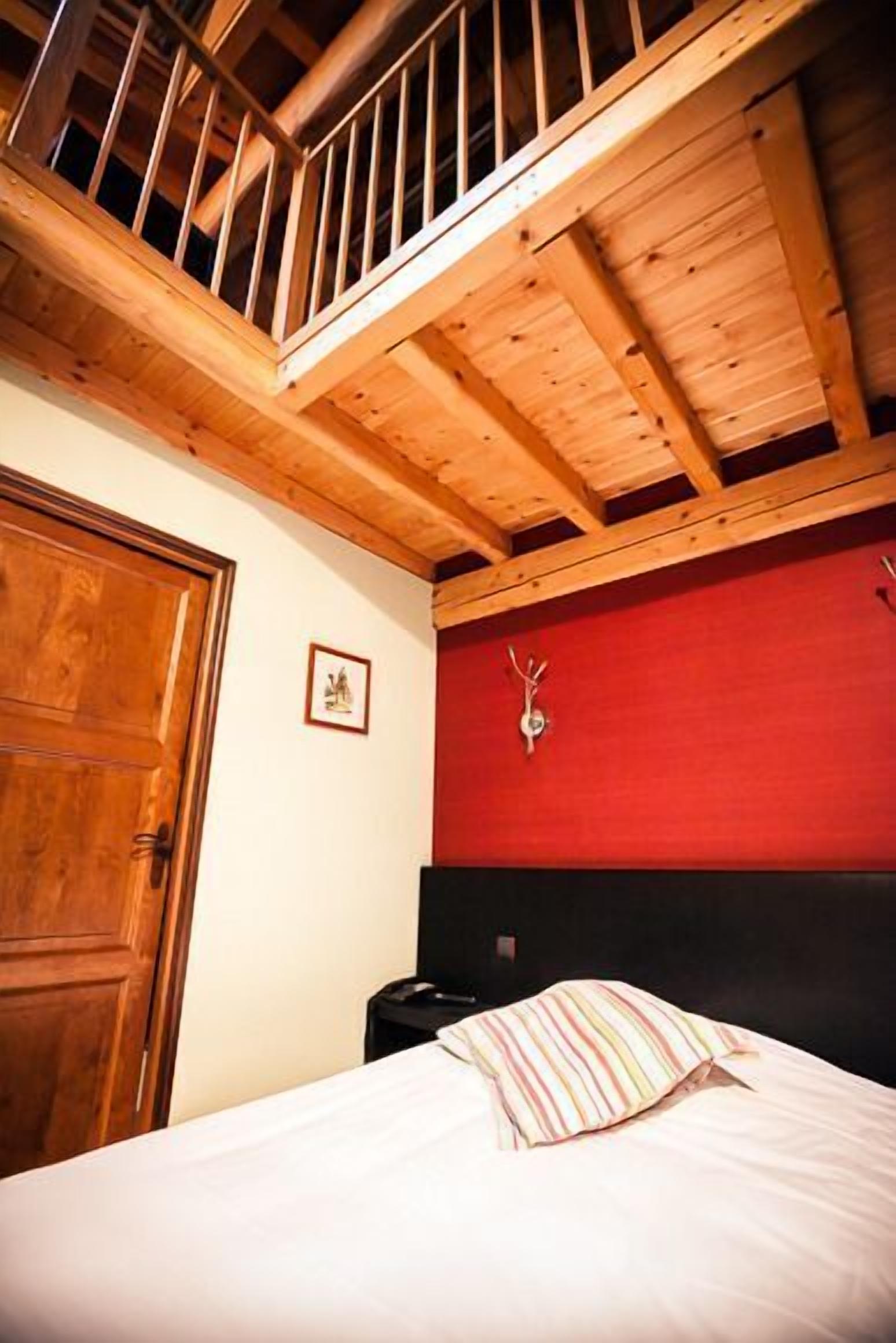 A bedroom with a wooden ceiling, a red accent wall, a bed with striped bedding, and a wooden door.