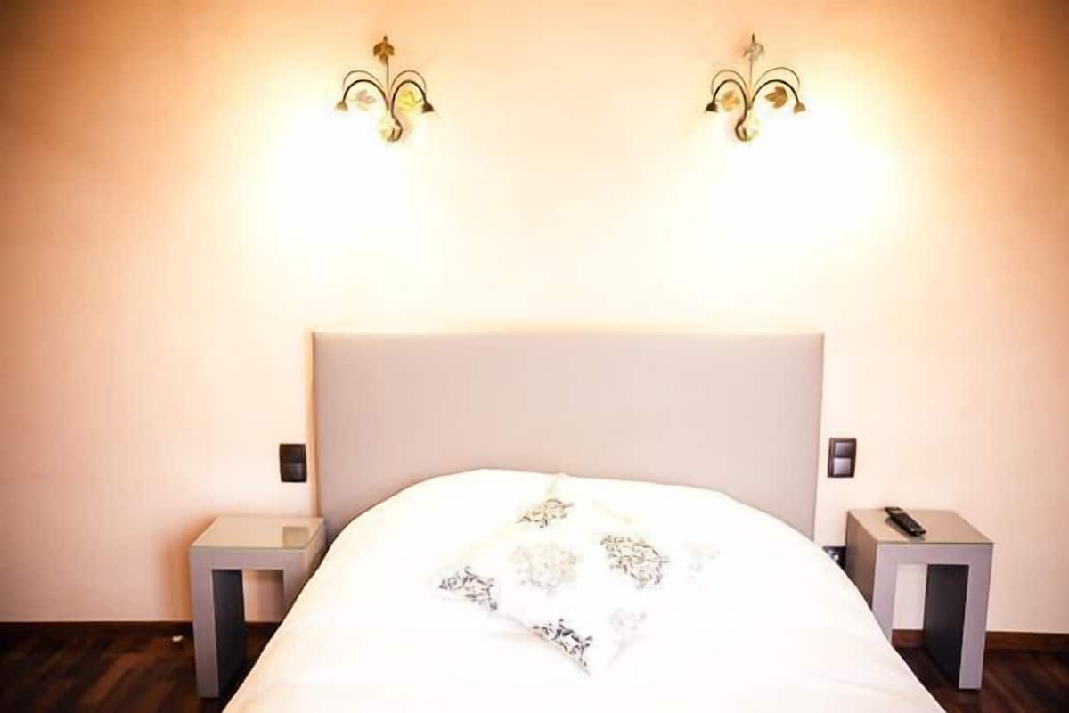 Una cama bien hecha con cabecera blanca y una manta con estampado floral. Hay dos mesitas de noche con lámparas a ambos lados de la cama.