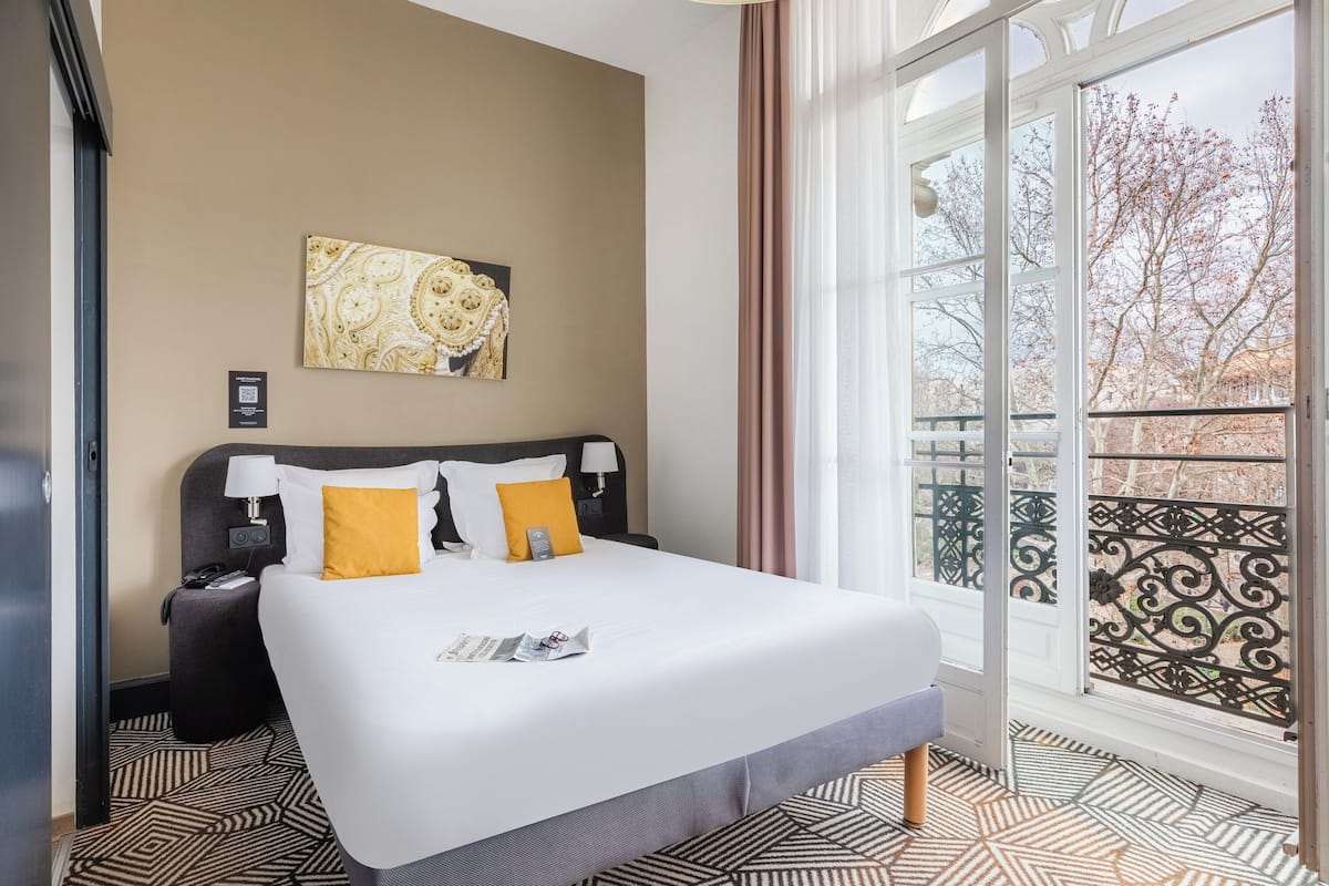 Ein Hotelzimmer mit einem großen Bett, einem Nachttisch, einer Lampe, einem gerahmten Kunstwerk an der Wand und Blick auf den Balkon.