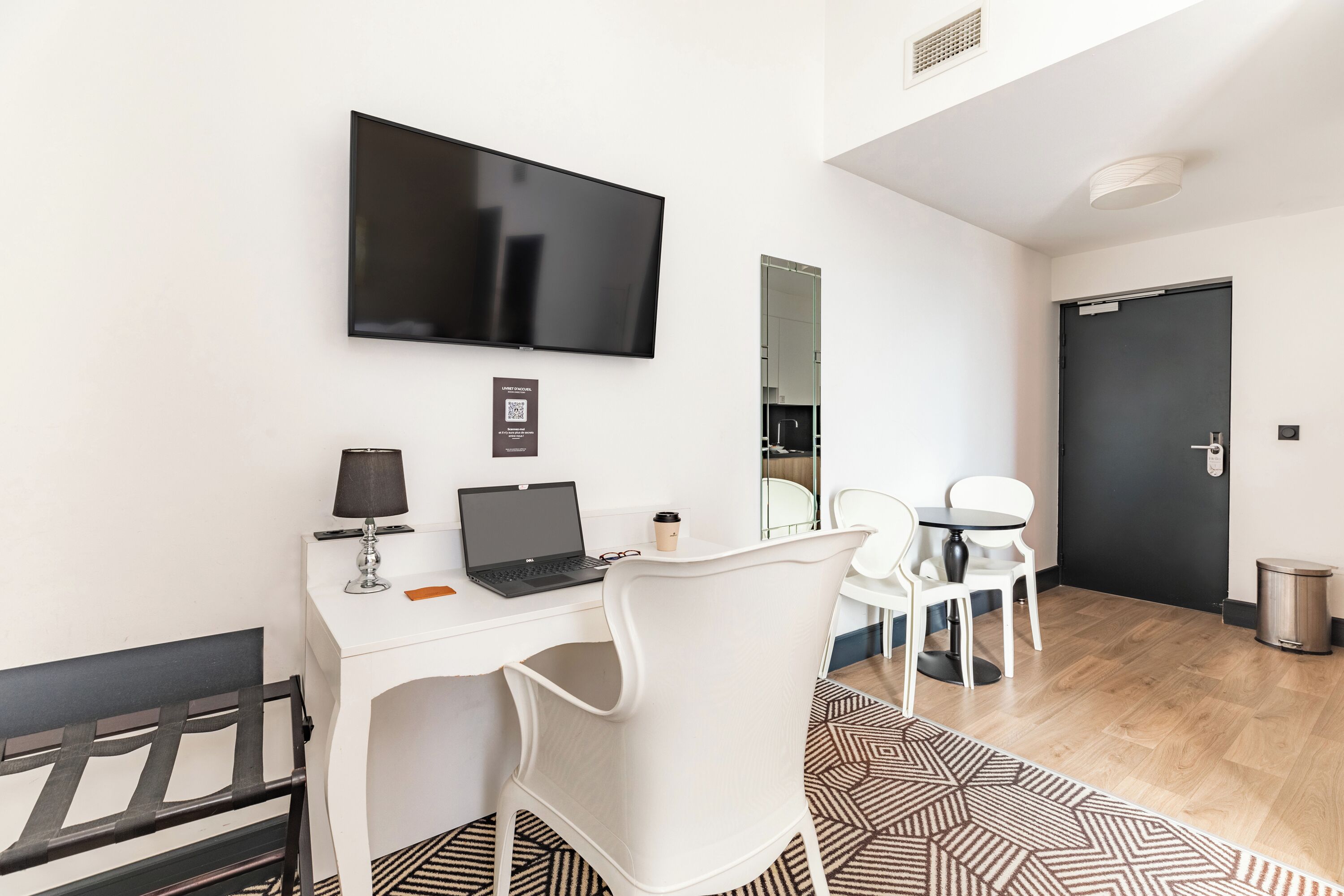 A modern hotel room with a white desk, a laptop, a flat-screen TV, a coffee cup, and a patterned rug.
