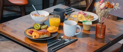 Una mesa de desayuno con un tazón de cereal, un vaso de jugo de naranja, una taza de café, un plato con croissants y un kiwi, y un florero con flores.
