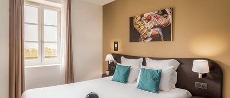 A hotel room with a large bed, two bedside tables with lamps, a window with curtains, and a painting on the wall.