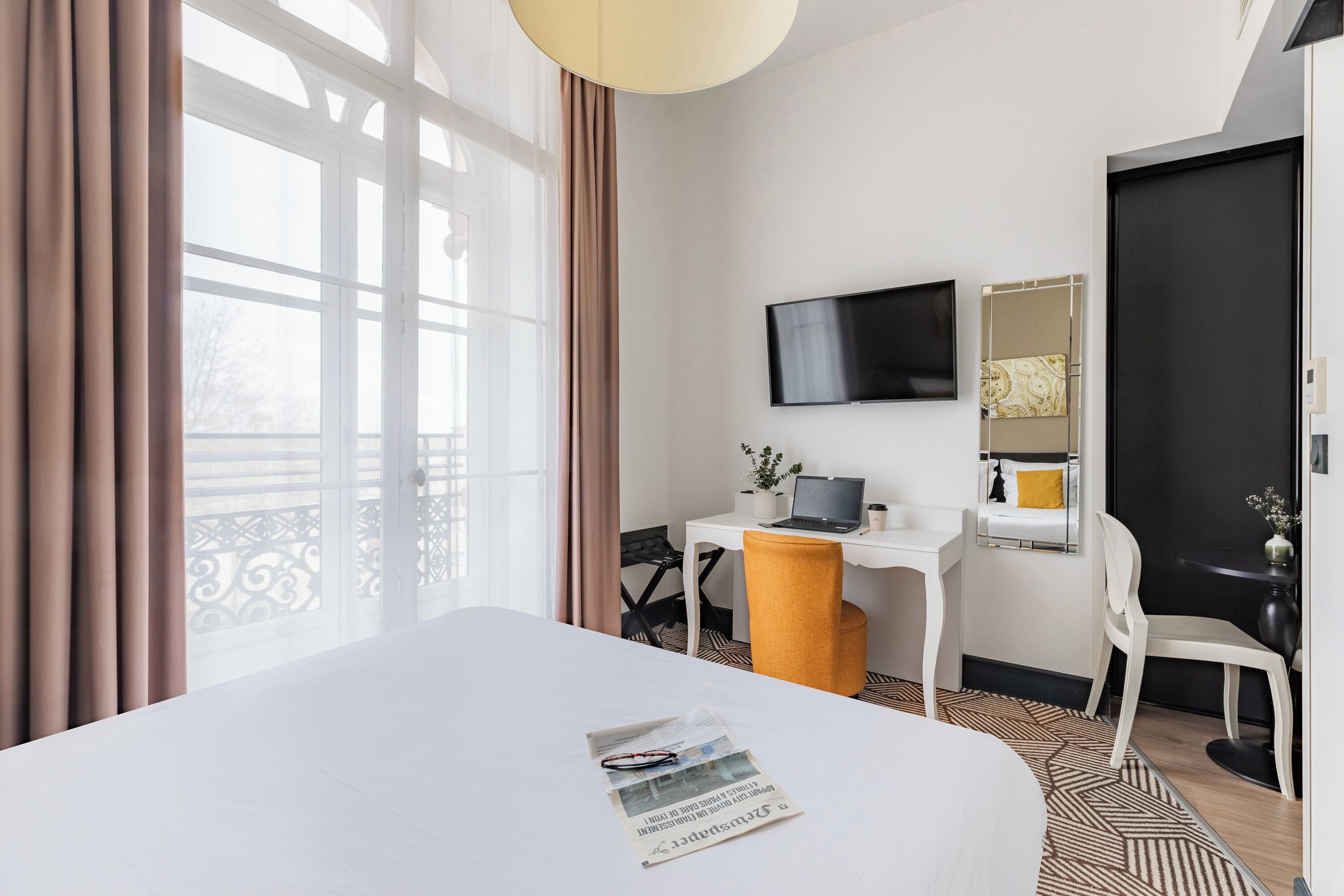 Ein Hotelzimmer mit einem großen Bett, einem Schreibtisch mit einem Laptop, einem an der Wand befestigten Fernseher und einem Fenster mit Vorhängen.