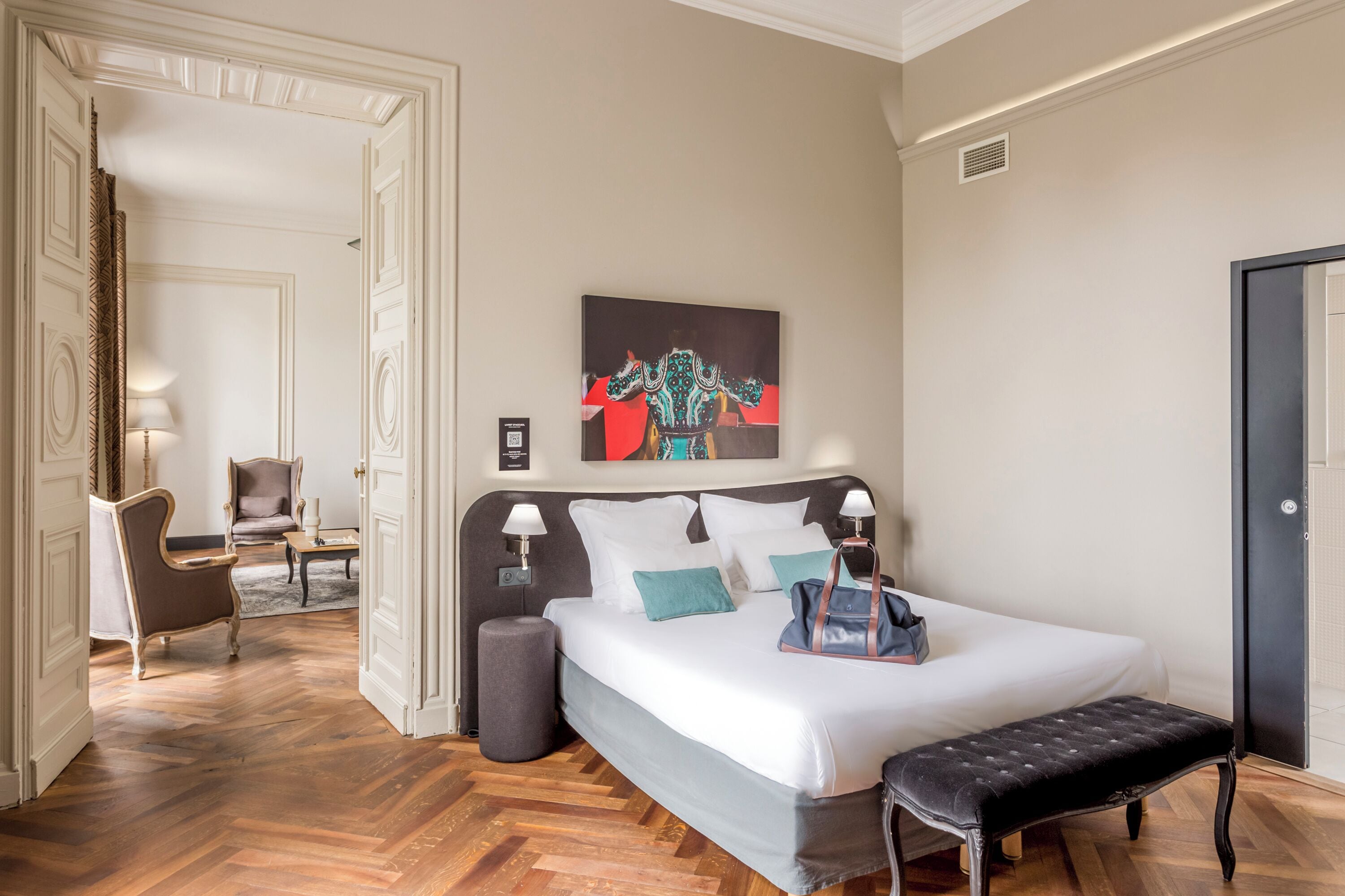 A hotel room with a large bed, a bench, a chair, and a painting on the wall.