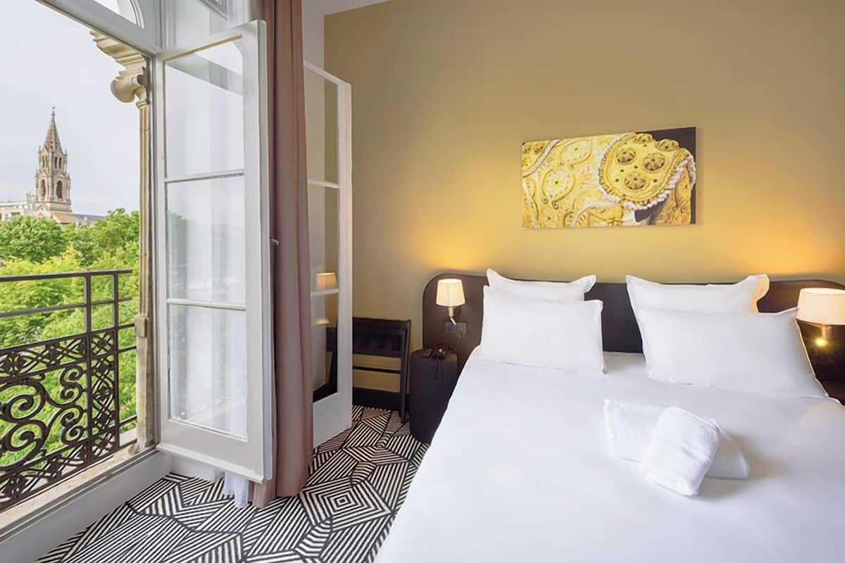 Ein Hotelzimmer mit einem großen Bett, einem Nachttisch, einer Lampe und Blick auf ein Gebäude durch ein Fenster.