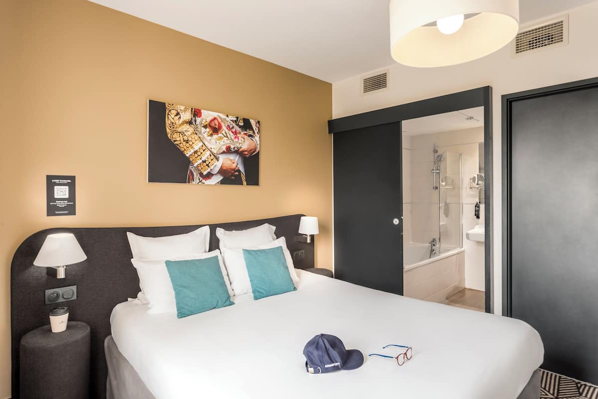 Ein Hotelzimmer mit einem großen Bett, zwei Nachttischen, einer Wandlampe, einem Gemälde und Blick ins Bad.