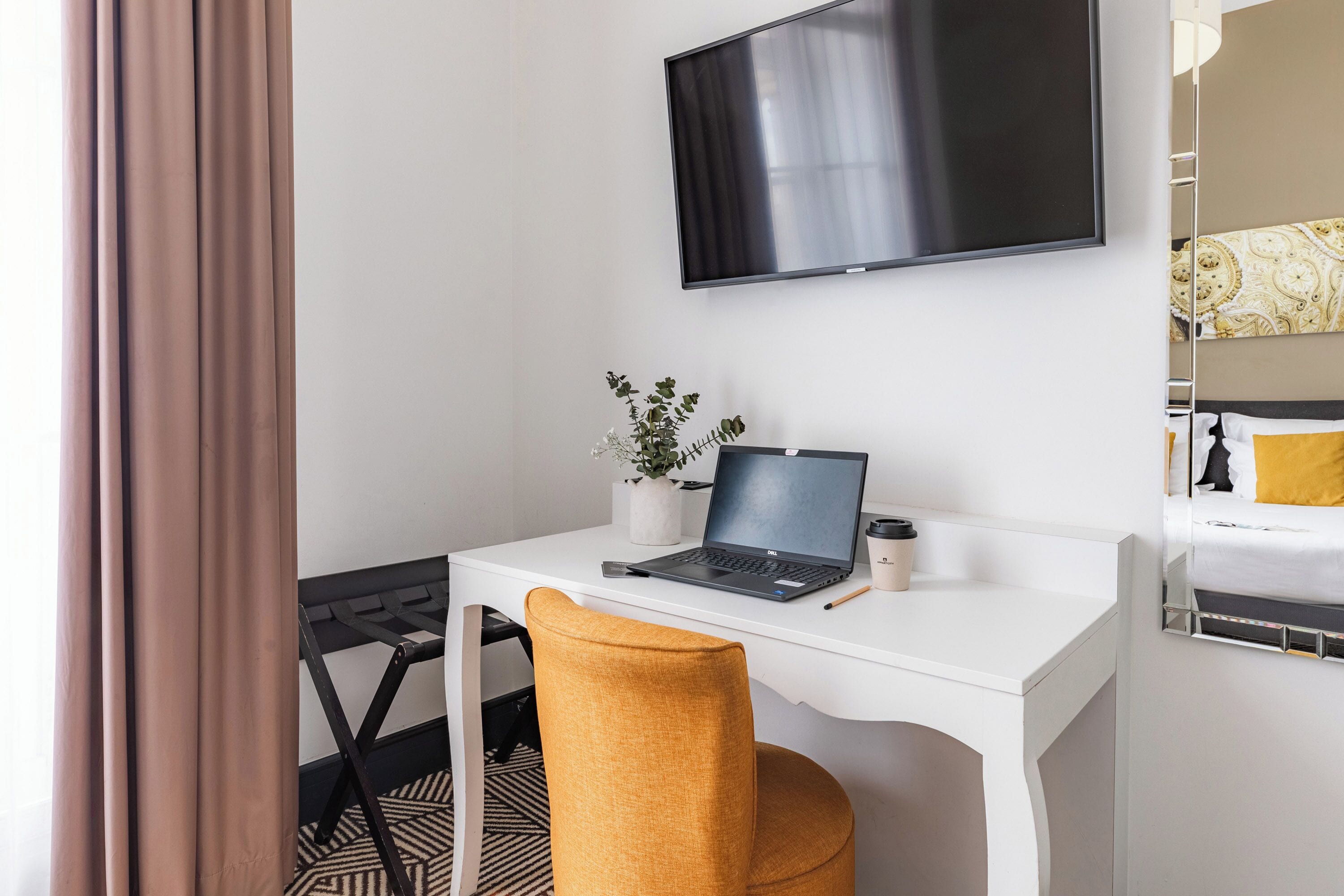 A hotel room with a white desk, a laptop, a small potted plant, and a bed with a patterned headboard.